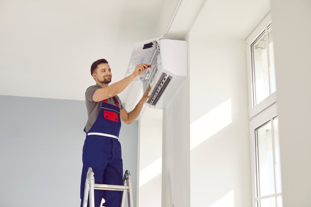 A technician in blue overalls stands on a ladder, repairing or installing an air conditioning unit on a white wall in a bright, modern room with large windows.