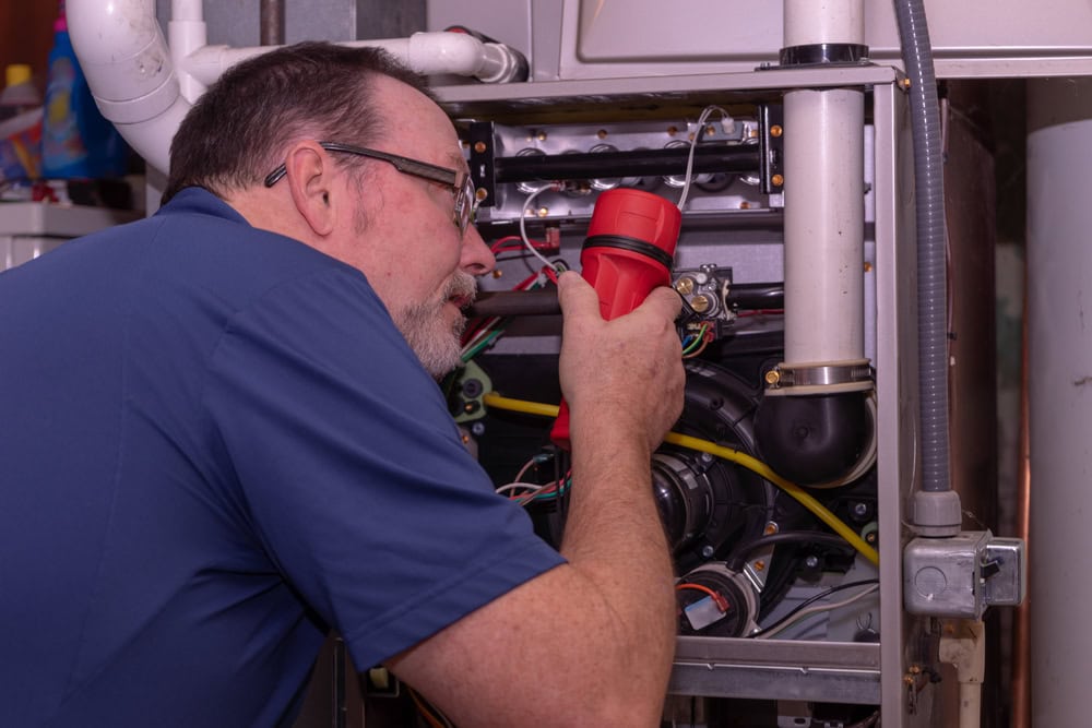 A man wearing glasses and a blue shirt uses a red flashlight to inspect the inside of a furnace, looking closely at the wiring and components.
