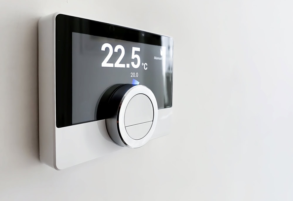 A modern digital thermostat mounted on a white wall displays a temperature of 22.5°C with a large round silver control dial beneath the screen.