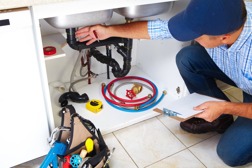A plumber in a blue cap inspects pipes under a kitchen sink, holding a clipboard. Plumbing tools, hoses, and a tool bag are visible on the floor and inside the open cabinet.