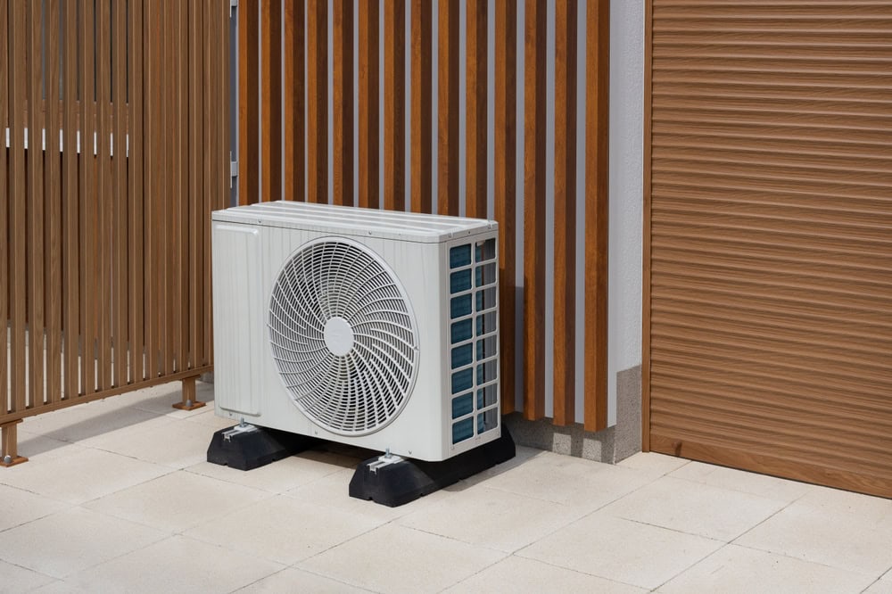 An outdoor air conditioning unit sits on a tiled patio next to a building with vertical wooden slats and a closed brown roller shutter door.