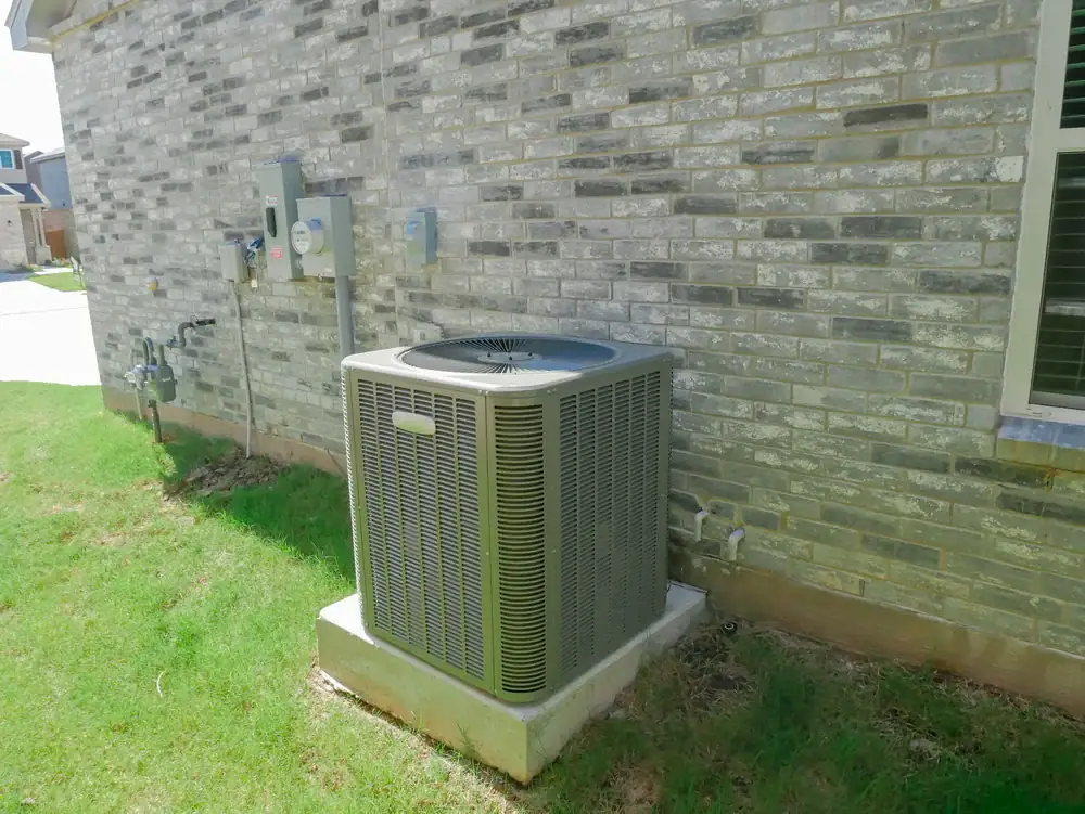 A gray outdoor air conditioning unit sits on a concrete pad by a brick house and green grass, highlighting expert AC installation North Attleboro homeowners trust. Electrical and utility boxes are mounted nearby on the wall.