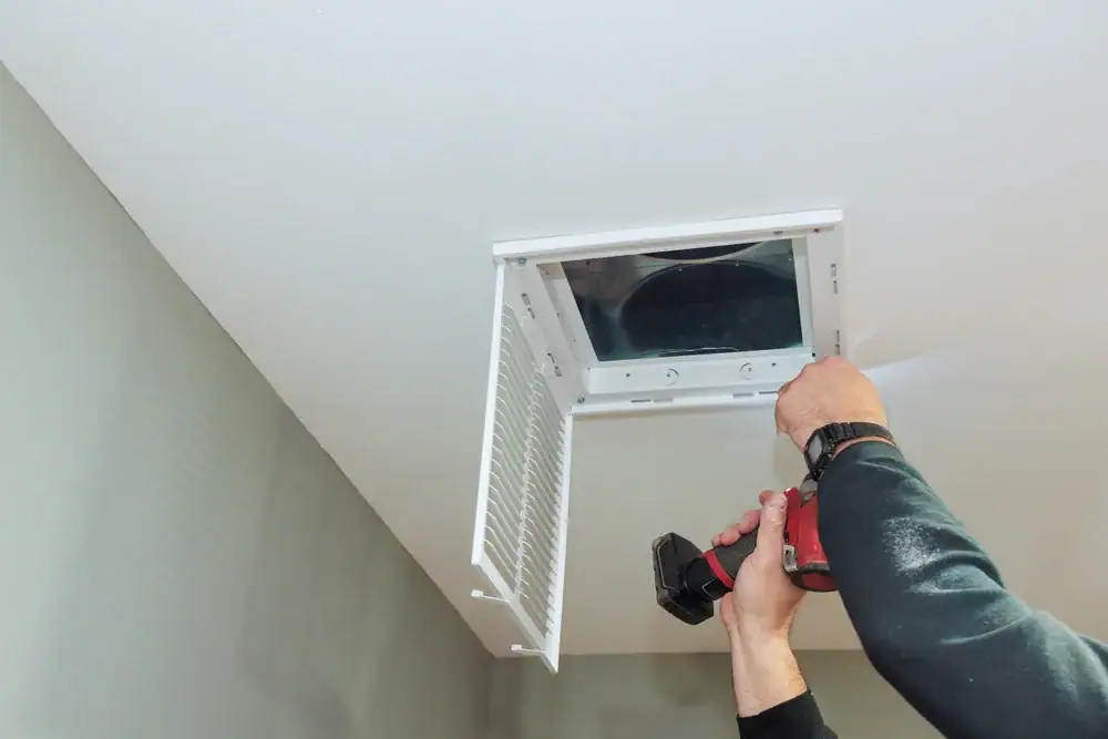 A person uses a power drill to install or remove an attic access panel in a white ceiling, with the door open and a dark attic space visible—ideal for AC Installation North Attleboro or when calling an HVAC Contractor North Attleboro.