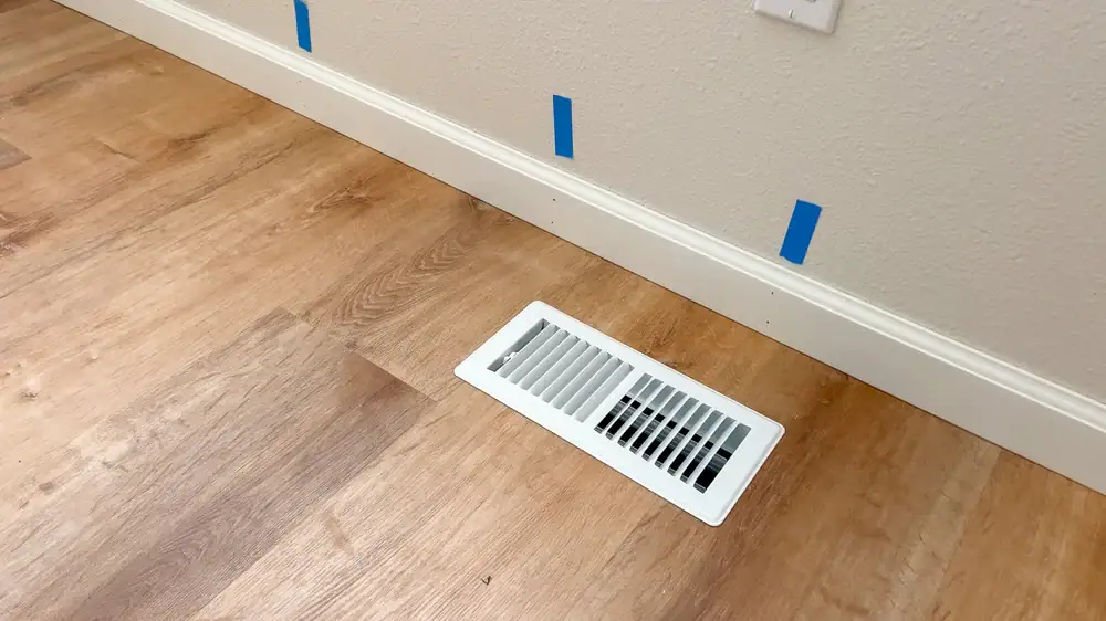 A white floor vent, installed on light wood flooring near a beige wall with a white baseboard, reflects the attention to detail you can expect from an expert HVAC Contractor in North Attleboro, MA. Blue painter’s tape marks line the baseboard evenly.