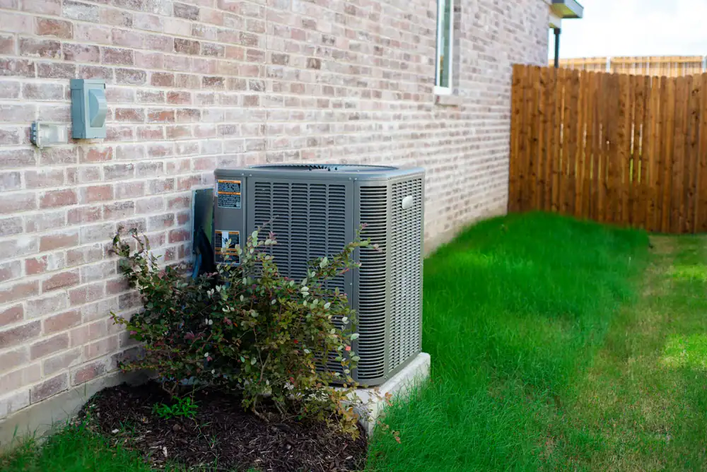 An outdoor air conditioning unit sits on a concrete pad next to a brick house in North Attleboro, MA, surrounded by green grass and a small bush&mdash;a typical scene for AC Installation North Attleboro and expert AC Services North Attleboro.
