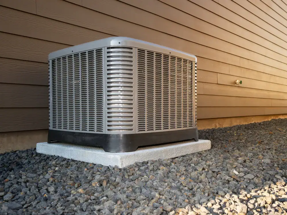 A central air conditioner unit sits on a concrete pad beside a house with brown siding, surrounded by gravel&mdash;expertly installed by an HVAC Contractor North Attleboro.
