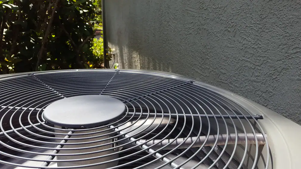 Close-up of the top of an outdoor air conditioning unit with a metal fan cover, next to a stucco-textured wall and partially shaded by nearby plants—perfect for highlighting AC Services North Attleboro, MA.