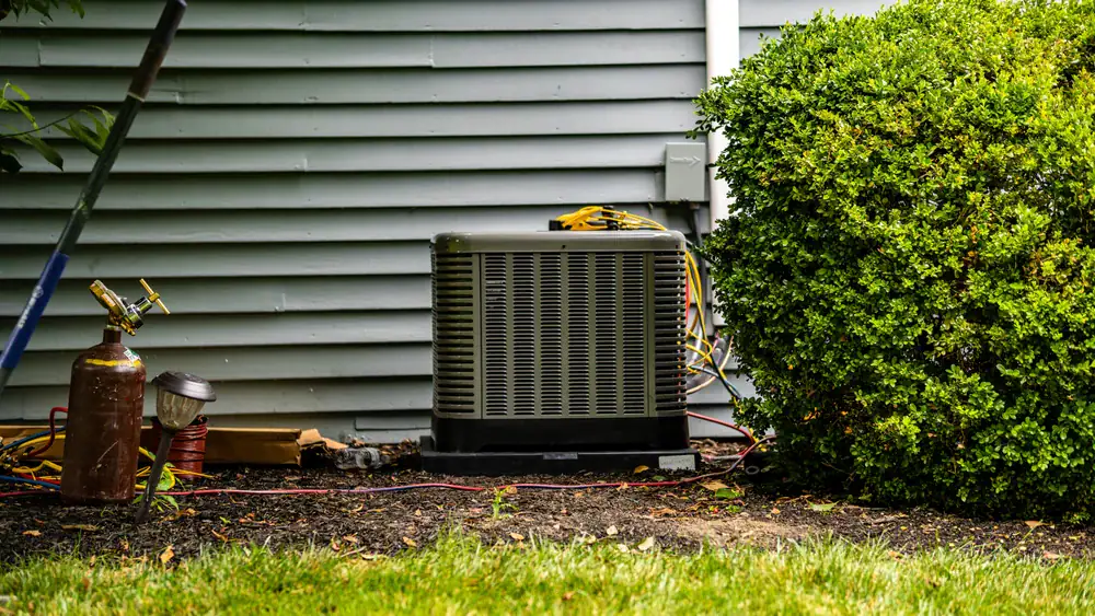 An outdoor air conditioning unit sits on the ground next to a house with gray siding, surrounded by bushes and grass, as an HVAC contractor North Attleboro prepares repair tools and equipment nearby.