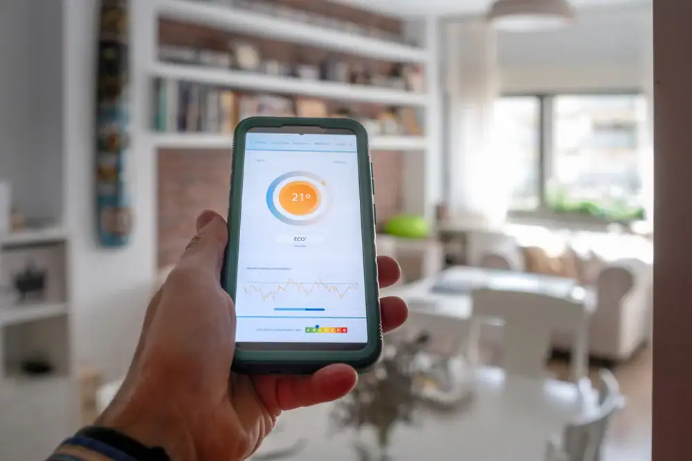 A person holds a smartphone displaying a home temperature app set to 21°C, with energy-saving data shown—perfect for those seeking expert AC Services North Attleboro in the bright, modern living room with shelves, table, and large windows.
