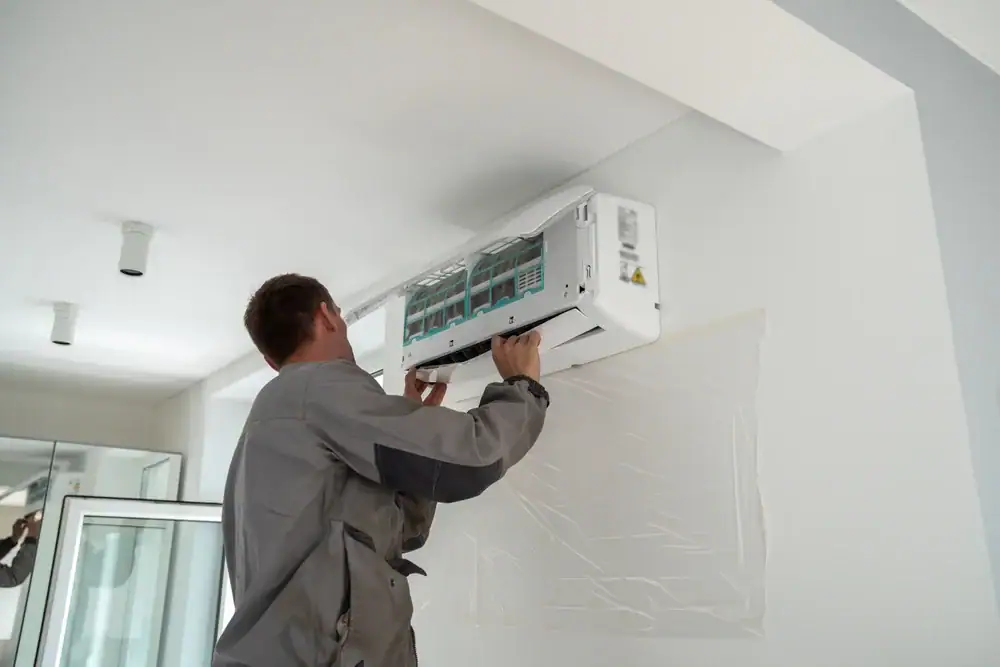A technician in a gray uniform is installing or servicing a wall-mounted air conditioning unit inside a modern, white room. As an HVAC Contractor North Attleboro, he works with both hands to ensure top-quality service.