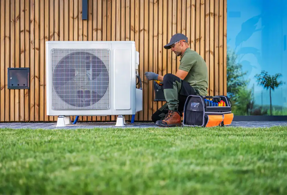 A technician wearing a cap and work gloves kneels on grass beside a house, servicing an outdoor AC unit with a toolbox nearby&mdash;your trusted HVAC Contractor North Attleboro for reliable service.