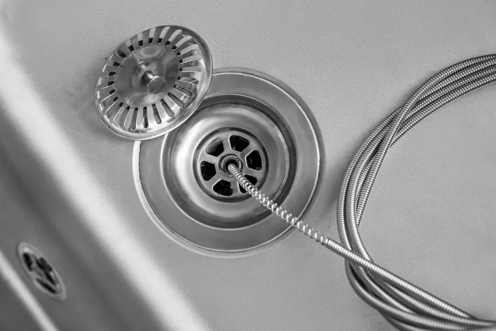 A close-up of a metal sink drain with a circular, slotted strainer partially lifted and a coiled flexible metal hose resting next to the drain on the smooth sink surface, often seen during AC Services North Attleboro, MA.