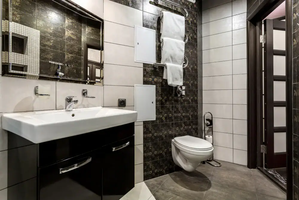 Modern bathroom with a wall-mounted toilet, rectangular sink with black cabinet, large mirror, stylish tiles, towel rack with folded white towels, and an open door—expertly maintained by your trusted HVAC Contractor North Attleboro, MA.