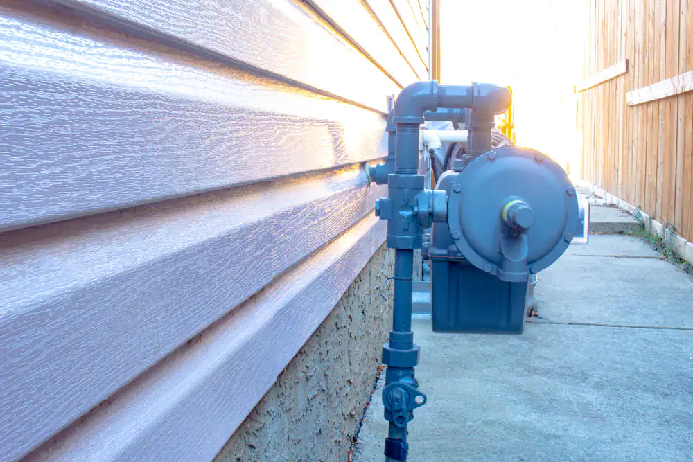 A natural gas meter and pipes are installed alongside the exterior wall of a house, next to a concrete pathway and wooden fence, in bright daylight&mdash;serviced by a trusted HVAC Contractor North Attleboro for reliable performance.