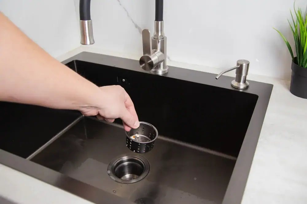 A hand places a metal strainer basket into the drain of a modern black kitchen sink with a silver faucet, soap dispenser, and a small green plant&mdash;perfect for homes serviced by an expert HVAC Contractor North Attleboro.