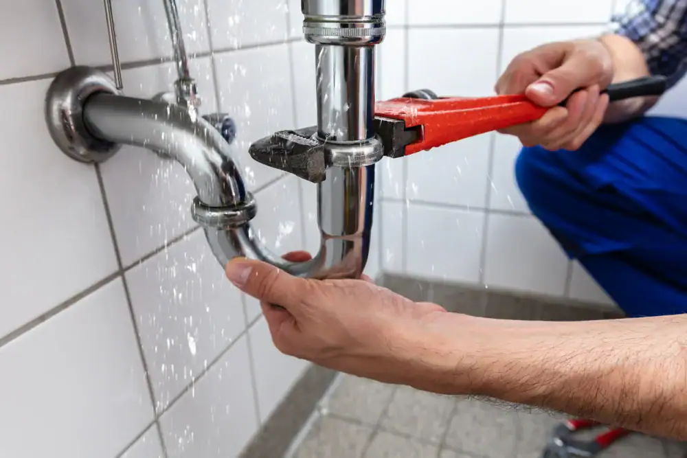 A person uses a red pipe wrench to fix a leaking metal pipe under a sink, with water spraying out onto tiled walls and floor—reminding homeowners in North Attleboro, MA, of the importance of timely plumbing and AC services.