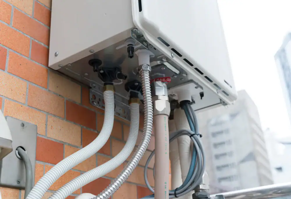 Close-up of pipes and cables connected to a white outdoor utility box mounted on a brick wall, with tall buildings in the blurred background&mdash;ideal setup for AC Installation North Attleboro, MA.