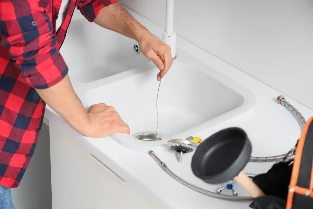 A person in a red plaid shirt uses a drain snake to unclog a white kitchen sink in North Attleboro, MA. Various plumbing and AC installation tools from AC Services North Attleboro are placed on the countertop nearby.
