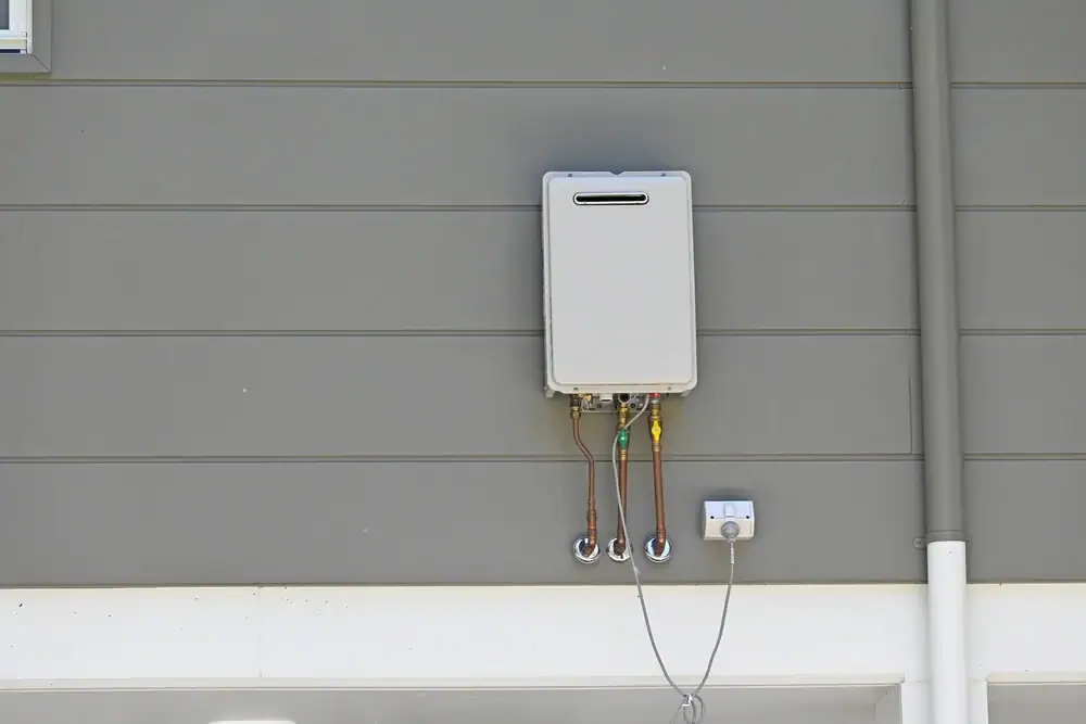 A tankless water heater is mounted on the exterior gray wall of a building, with several pipes and wiring connected beneath it—professionally installed by an HVAC Contractor North Attleboro.