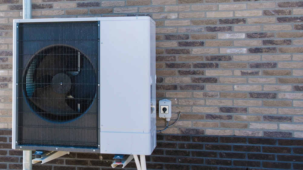 A white outdoor air conditioning unit is mounted on a brick wall in MA. The unit features a large circular fan behind a protective grille and connects to a nearby outlet, showcasing quality AC Installation North Attleboro residents can trust.