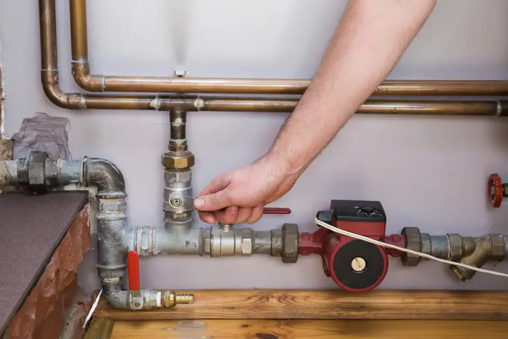 A person’s hand turning a valve on a pipe system attached to a wall, with copper and metal pipes visible, including a red water pump and gauges—ideal for showcasing quality AC Installation in North Attleboro by an expert HVAC contractor.