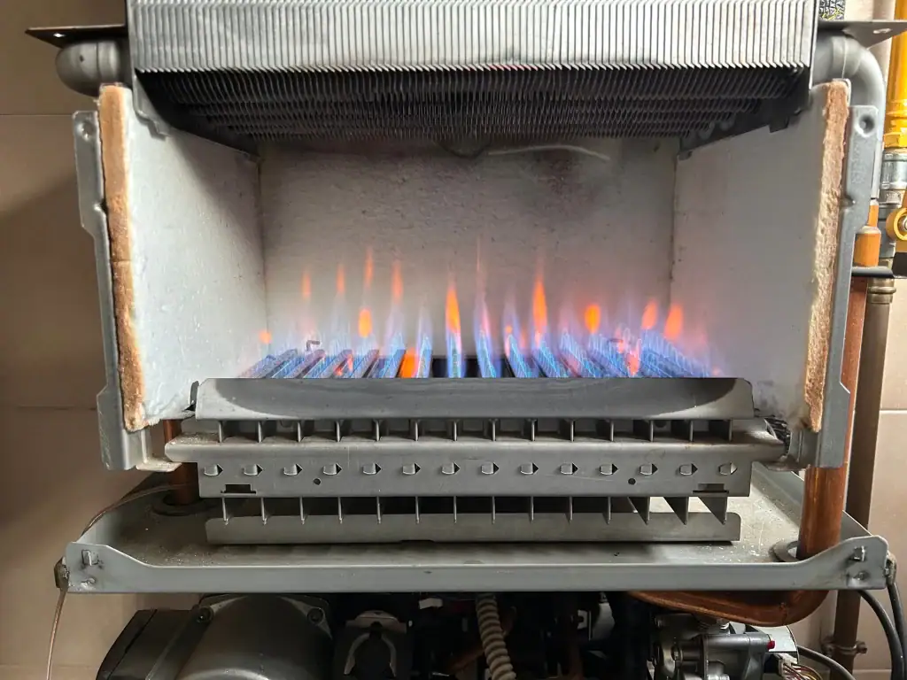 A close-up of a gas boiler in operation, showing blue and orange flames heating metal pipes inside the unit. The metallic interior and insulation panels highlight quality, similar to top AC Installation North Attleboro services in MA.
