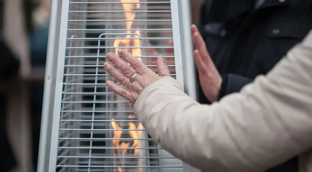 A person in a light-colored coat warms their hands near a tall outdoor heater—perfect for chilly evenings when AC Services North Attleboro, MA, or an HVAC Contractor North Attleboro is needed to keep the indoors comfortable. Another person stands nearby, also warming up.
