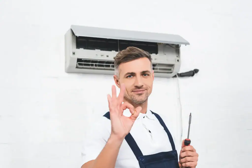 A technician in overalls holding a screwdriver stands in front of an open wall-mounted air conditioner, smiling and making an "OK" hand gesture.